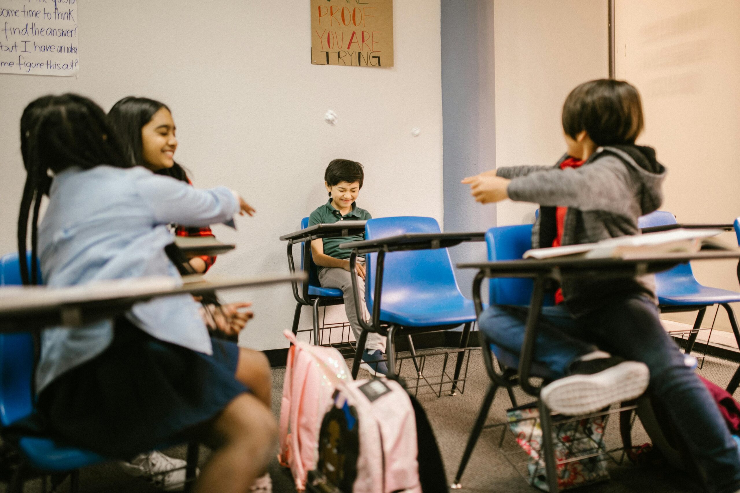 Four children in classroom-style desks. Two are throwing something at a third child, who is squinting at the nearing objects